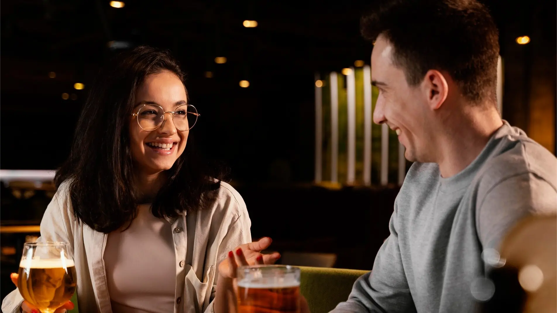 Junges Paar beim ersten Date in gemütlicher Bar – Frau mit Brille und Mann im grauen Pullover lachen und genießen Bier – authentischer Moment voller Sympathie und Verbindung